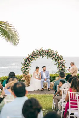 Pareja sentada frente a un arco floral circular durante su ceremonia de Destination Wedding con vista al océano.