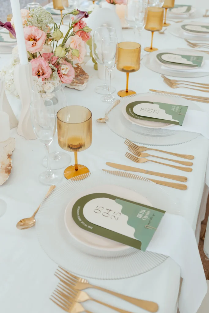 Vajilla con cristalería ámbar y papelería personalizada en tonos verdes para un banquete de matrimonio igualitario.